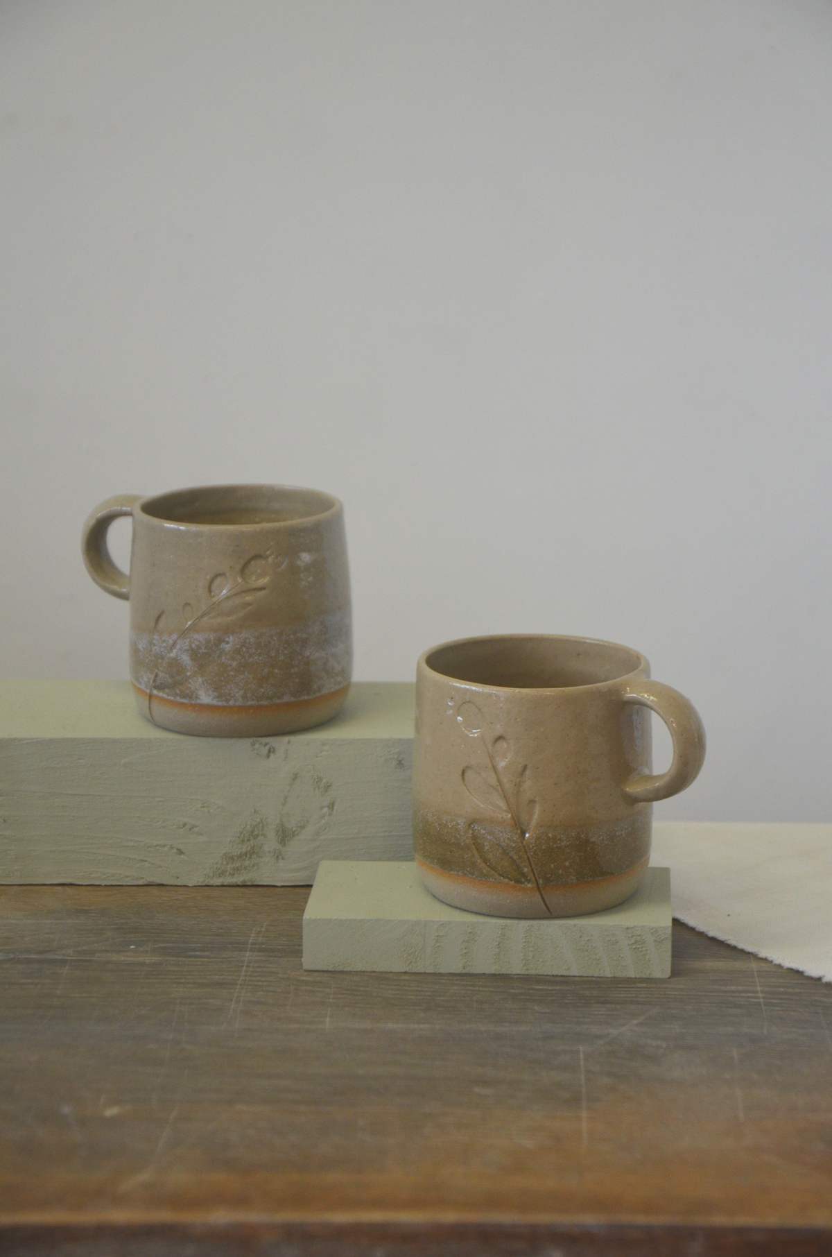 Deux tasses beiges à anse, décor de brins végétaux, posées sur des socles.